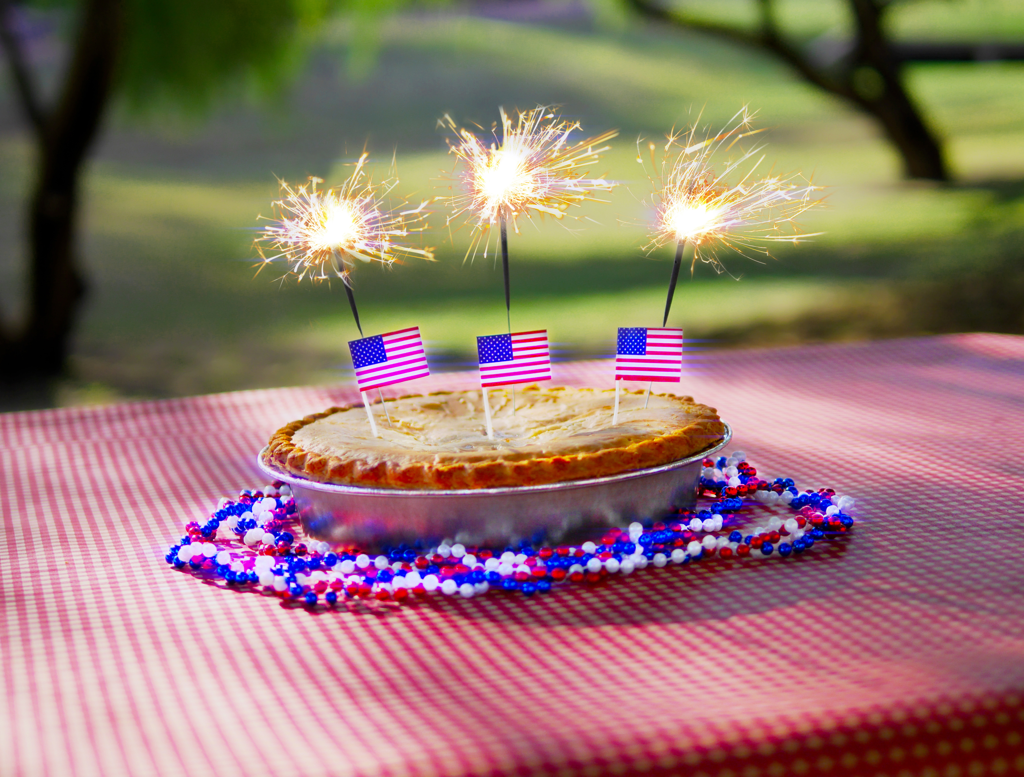 A Fourth of July apple pie with red, white, and blue beads, American flags, and sparklers on a picnic table in a park.